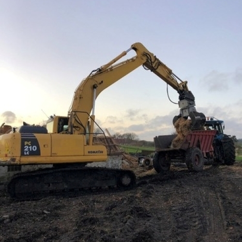 En Komatsu gravemaskine læsser jord og murbrokker op i en rød traktoranhænger på en mudret markvej. Nedrivningsarbejdet foregår i aftensol.