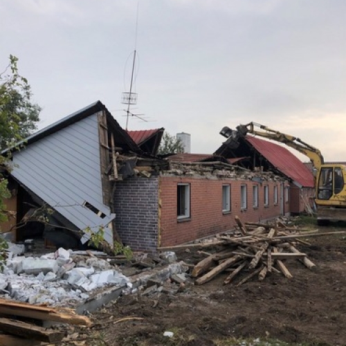 En gul gravemaskine river et langt, lavt hus ned. Træ, mursten og bygningsdele ligger i bunker foran den delvist kollapsede bygning.