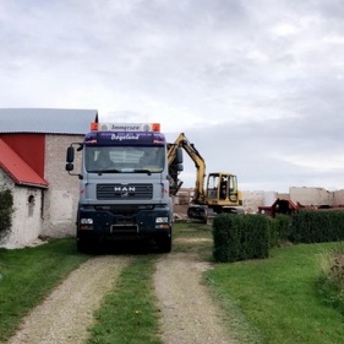 En lastbil og en gravemaskine arbejder sammen ved et hus på landet. Lastbilen holder på en grusvej, mens gravemaskinen fjerner bygningsdele i baggrunden.