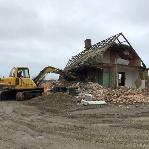 En gul gravemaskine nedbryder væggen på et hus. Bygningen er delvist revet ned, og mursten og træ ligger i bunker foran maskinen.