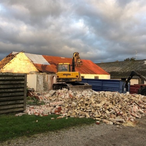 En gravemaskine river en murstensbygning ned under aftensol. Foran huset ligger store mængder af nedrevne mursten klar til bortkørsel.