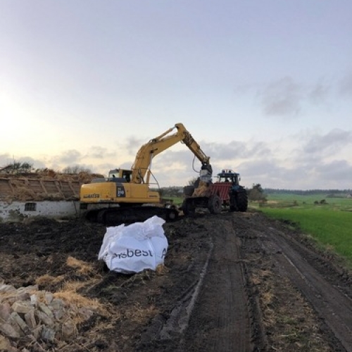 En gravemaskine rydder op efter nedrivning af en bygning i landlige omgivelser. En traktor med anhænger hjælper med at fjerne murbrokker fra pladsen.