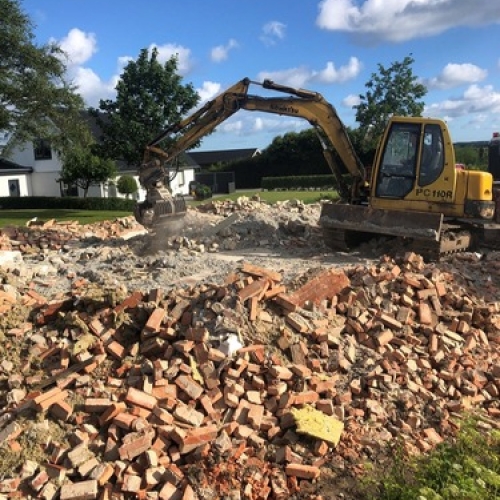 En gravemaskine rydder murbrokker og betonrester efter en nedrivning. Arbejdet foregår på en solrig dag med blå himmel og grønne omgivelser.