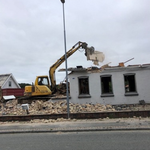 En gul gravemaskine river taget af en hvid bygning. Murbrokker og tagdele ligger spredt omkring huset, mens arbejdet fortsætter.