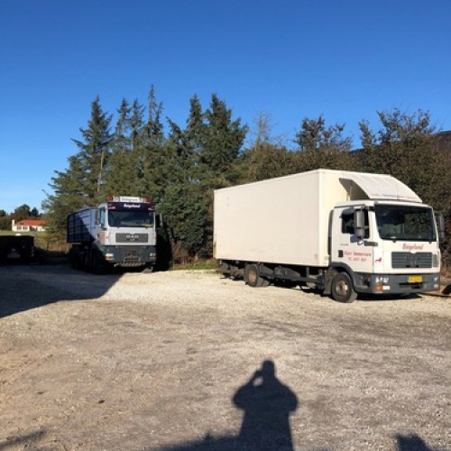 To lastbiler fra samme entreprenørfirma holder parkeret foran en række træer på en solrig dag. Den ene lastbil er med container, den anden en lukket flyttevogn.
