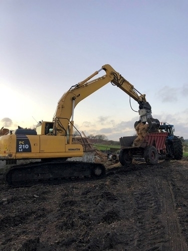 En Komatsu gravemaskine læsser jord og murbrokker op i en rød traktoranhænger på en mudret markvej. Nedrivningsarbejdet foregår i aftensol.