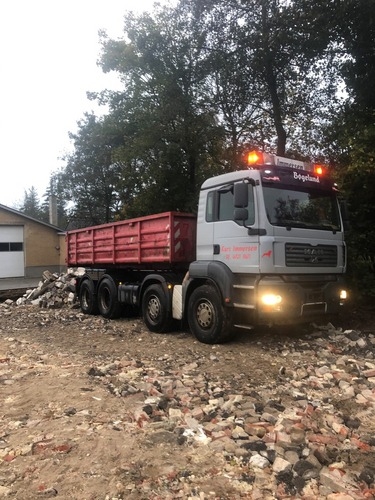 En tung lastbil med tændte advarselslys holder på en byggeplads omgivet af murbrokker og sten. Lastbilen er klar til bortkørsel af byggeaffald efter nedrivning.