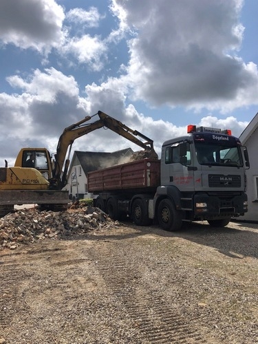 En gravemaskine læsser murbrokker og byggeaffald op i en rød lastbil på en solrig dag. Arbejdet foregår på en byggeplads mellem huse.