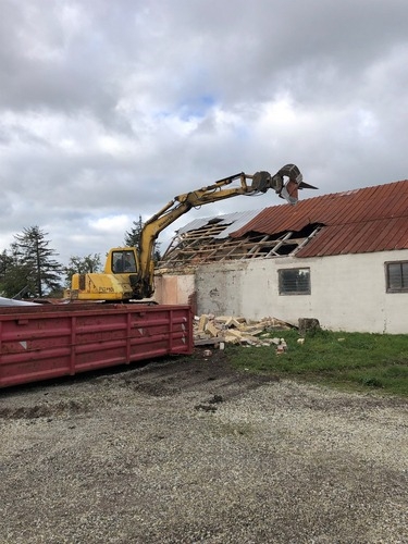 En gul gravemaskine fjerner taget fra en ældre bygning. Tagplader og træværk bliver revet ned som en del af en nedrivningsopgave.