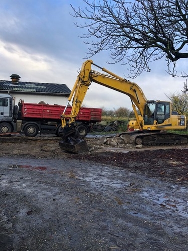 En gravemaskine graver og læsser jord eller byggemateriale op i en rød lastbil på en byggeplads. Omgivelserne er våde og mudrede efter regn.