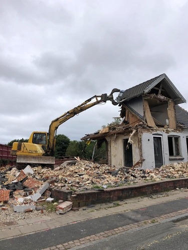 En gravemaskine nedbryder et to-etagers hus af mursten. Bygningen er delvist revet ned, og murbrokker ligger spredt foran huset.