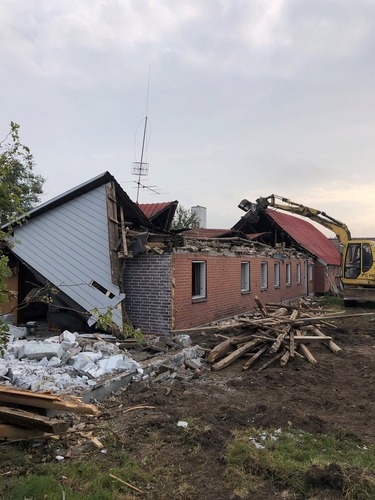 En gul gravemaskine river et langt, lavt hus ned. Træ, mursten og bygningsdele ligger i bunker foran den delvist kollapsede bygning.