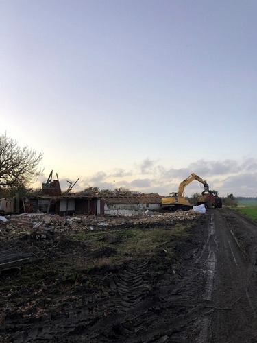 En gravemaskine arbejder på at rydde resterne af en nedrevet bygning i landlige omgivelser. Jordvejen foran er præget af dybe hjulspor.