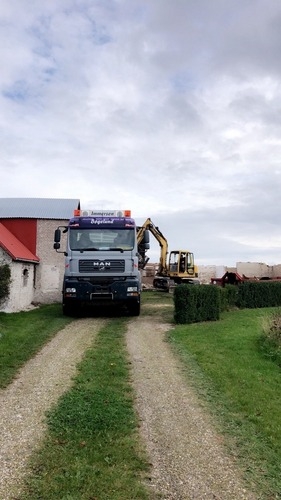 En lastbil og en gravemaskine arbejder sammen ved et hus på landet. Lastbilen holder på en grusvej, mens gravemaskinen fjerner bygningsdele i baggrunden.