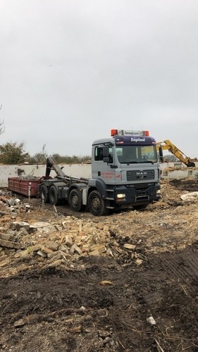 En lastbil med lad til byggeaffald holder på en byggeplads omgivet af murbrokker og jord. En gravemaskine arbejder bagved med at rydde området.