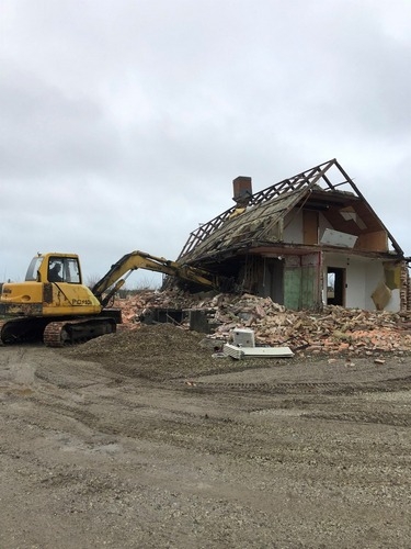 En gul gravemaskine nedbryder væggen på et hus. Bygningen er delvist revet ned, og mursten og træ ligger i bunker foran maskinen.