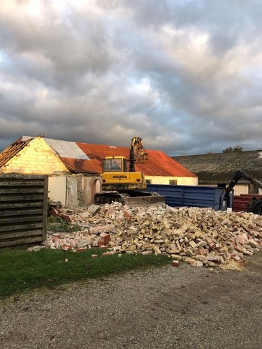 En gravemaskine river en murstensbygning ned under aftensol. Foran huset ligger store mængder af nedrevne mursten klar til bortkørsel.