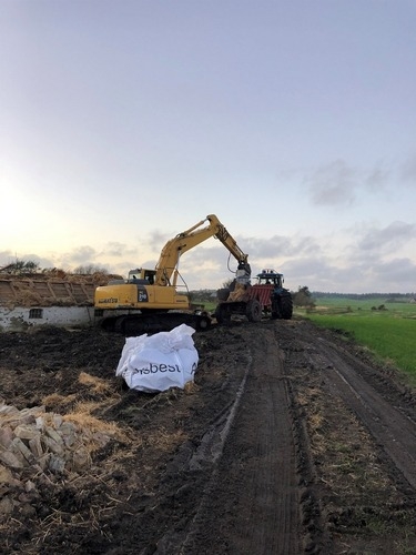 En gravemaskine rydder op efter nedrivning af en bygning i landlige omgivelser. En traktor med anhænger hjælper med at fjerne murbrokker fra pladsen.