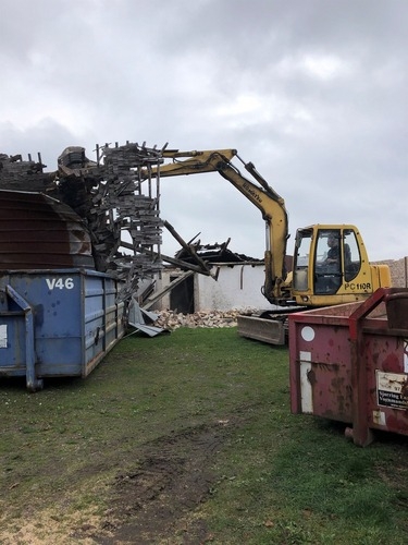 En gravemaskine fjerner tagkonstruktionen fra en ældre bygning. Træbjælker og tagmateriale falder ned i containere under arbejdet.