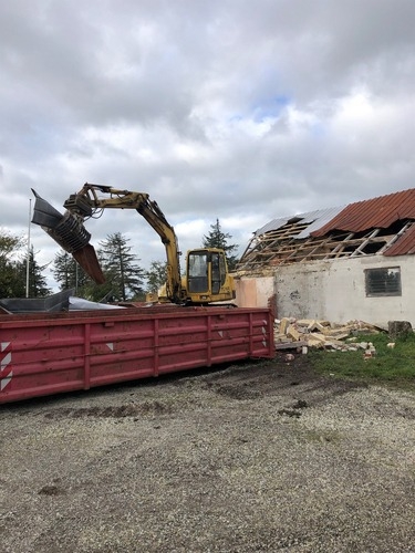 En gravemaskine fjerner taget på en ældre bygning. Metaldelene fra taget bliver lagt i en rød container ved siden af bygningen under nedrivningen