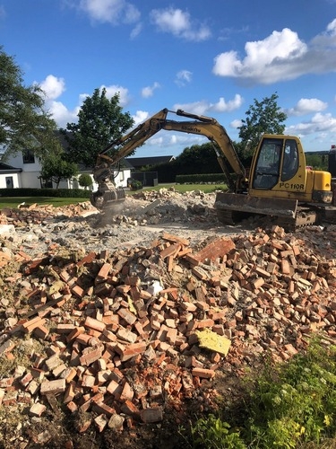 En gravemaskine rydder murbrokker og betonrester efter en nedrivning. Arbejdet foregår på en solrig dag med blå himmel og grønne omgivelser.