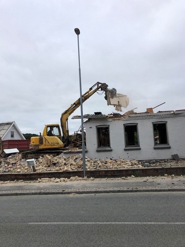 En gul gravemaskine river taget af en hvid bygning. Murbrokker og tagdele ligger spredt omkring huset, mens arbejdet fortsætter.