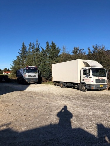 To lastbiler fra samme entreprenørfirma holder parkeret foran en række træer på en solrig dag. Den ene lastbil er med container, den anden en lukket flyttevogn.